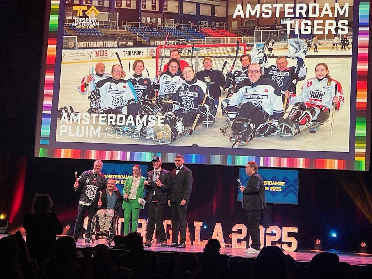 Op een groot podium staan 4 vertegenwoordigers van de Amsterdam Tigers naast wethouder Sport Mbarki. Een van hen heeft het beeldje van de Pluim in handen. Op eem groot scherm zien we een groepfoto van de ijshockeyers op de ijsbaan in hun uitrusting op de slee.