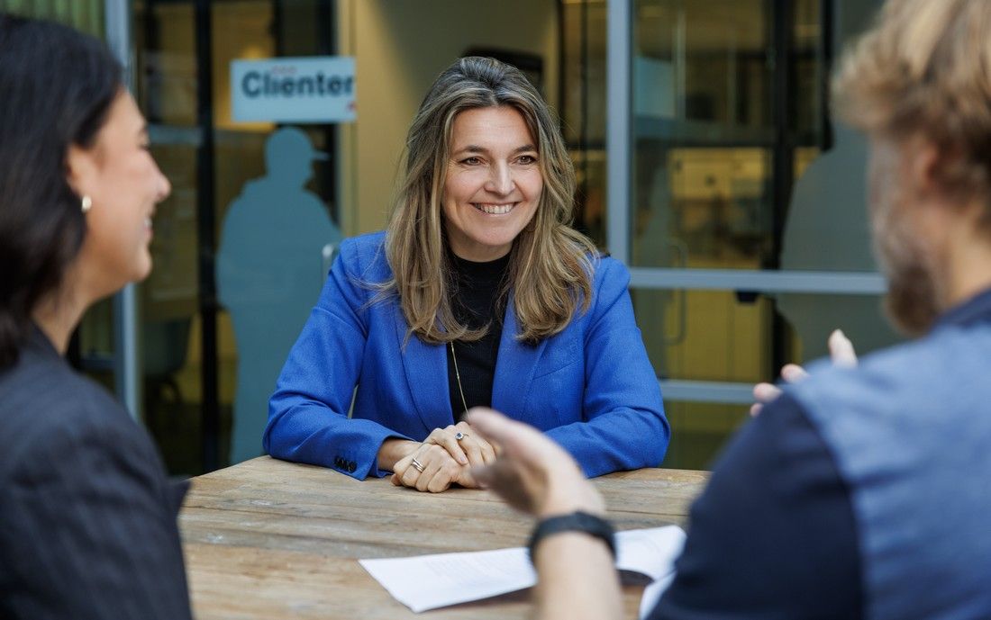 Lindy van Vliet in gesprek met 2 mensen aan een tafel in de grote hal bij de ingang van het kantoor van Cliëntenbelang Amsterdam