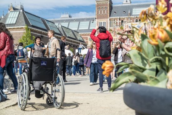 Groep mensen op Museumplein met het Rijksmuseum op de achtergrond en een rolstoel met een jas op de zitting op de voorgrond.